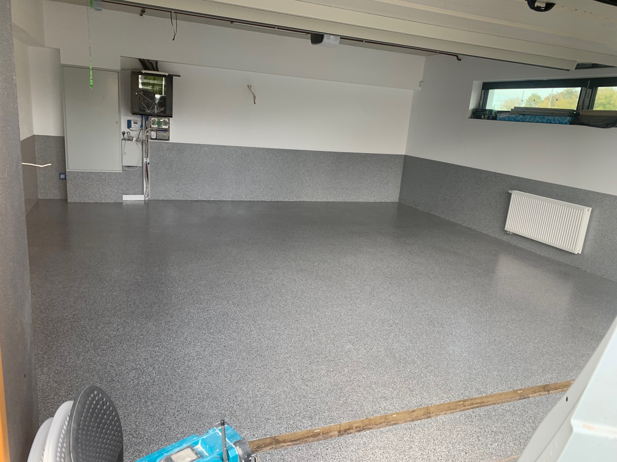 Szara, gotowa posadzka z drobnymi plamkami w garażu, z grzejnikiem na ścianie i niebieskim urządzeniem do czyszczenia podłóg w rogu.