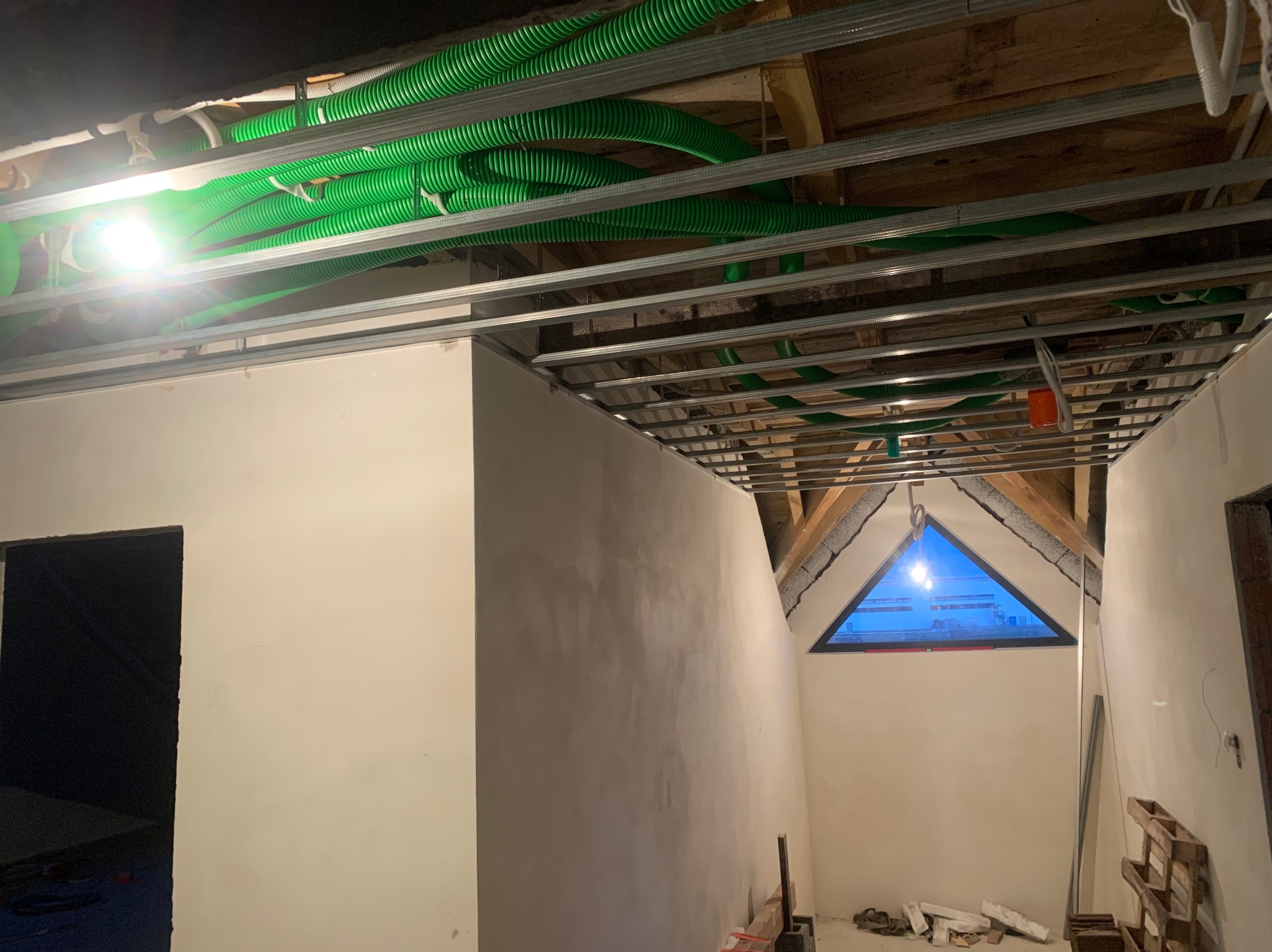 Wnętrze poddasza w trakcie prac wykończeniowych, widoczne metalowe profile sufitowe z instalacją elektryczną w zielonych rurach karbowanych, ściany po szpachlowaniu, trójkątne okno na szczycie...