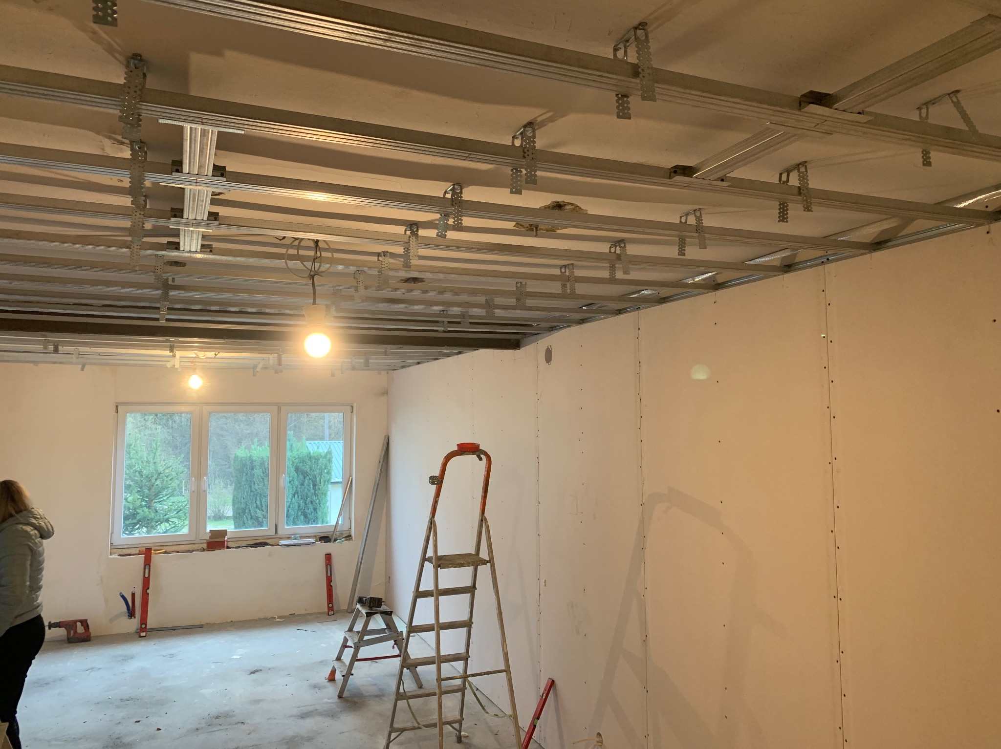 Instalacja stelaża sufitu podwieszanego z płyt karton gips w trakcie montażu, widoczne profile metalowe, mocowania, drabiny i częściowo zamontowane płyty na ścianach, okno z widokiem na zieleń.