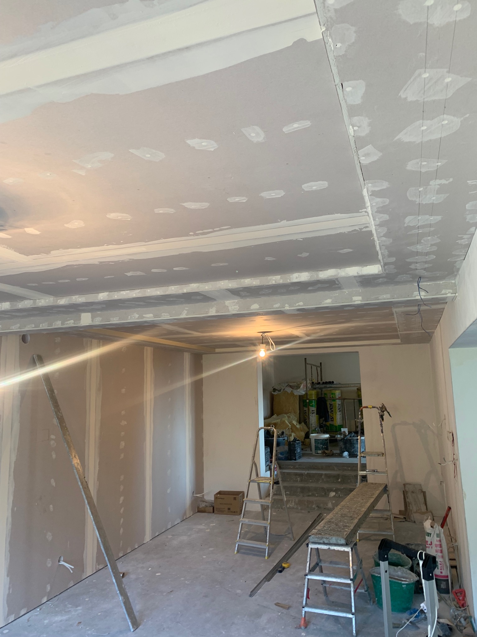 Wnętrze w trakcie remontu: ściany wyłożone płytami gipsowo-kartonowymi, sufit z elementami szpachlowania, widoczne drabiny i narzędzia budowlane, przygotowania do dalszych prac wykończeniowych.
