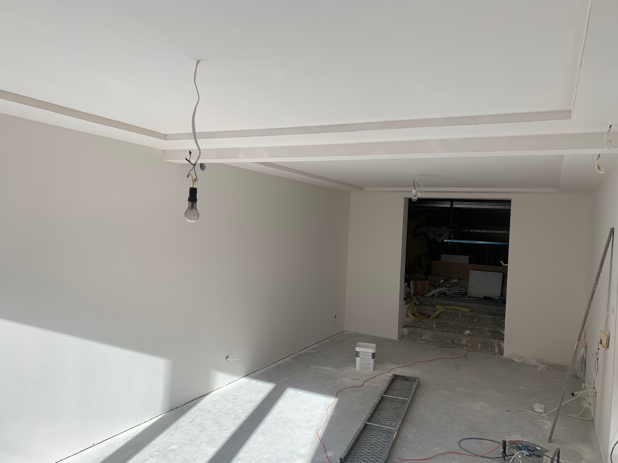Wnętrze w trakcie prac wykończeniowych: świeżo pomalowane ściany w odcieniach bieli, sufit z dekoracyjnymi elementami, wisząca żarówka na kablu, betonowa podłoga w trakcie przygotowania, widoczne...