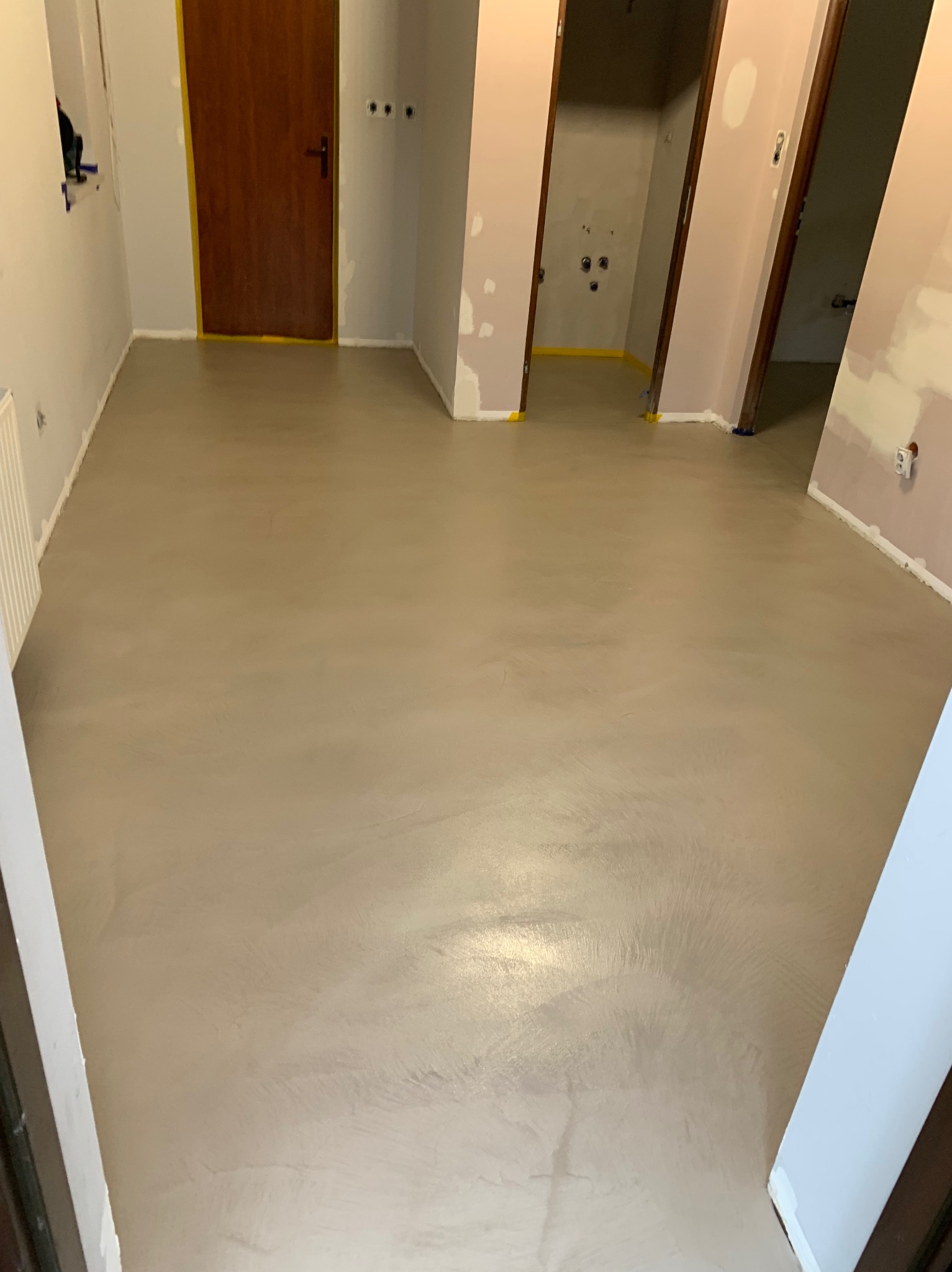 Jasnoszara, gładka posadzka w trakcie prac wykończeniowych, widoczne zabezpieczone taśmą malarską ościeżnice drzwiowe i ściany w surowym stanie.