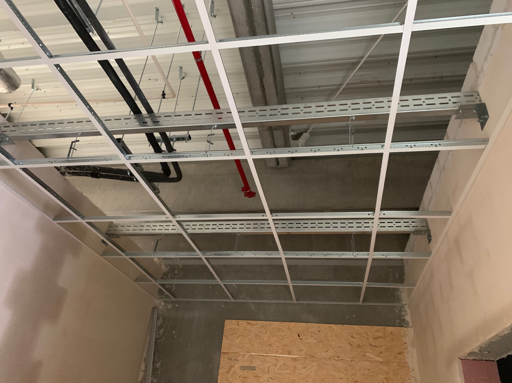 Widok z dołu na metalowy stelaż sufitowy z widocznymi instalacjami: rurami i kablami, zamontowany w surowym wnętrzu z fragmentami ścian po szpachlowaniu i płytą OSB przy podłodze.