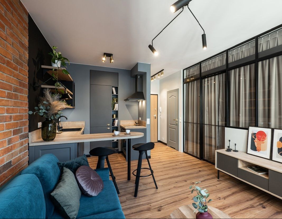 Salon z aneksem kuchennym w stylu industrialnym: ceglana ściana, szara zabudowa kuchenna, drewniany blat baru, turkusowa sofa z poduszkami, podłoga z paneli, przeszklenie w czarnej ramie z zasłonami.