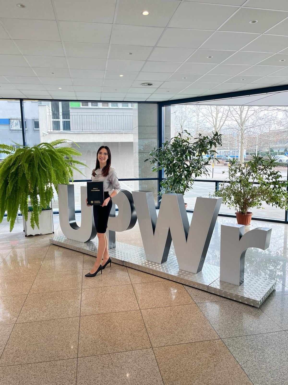 Kobieta w szarej bluzce i czarnej spódnicy pozuje z dyplomem na tle dużych liter tworzących skrót nazwy uczelni, z roślinami doniczkowymi w tle.