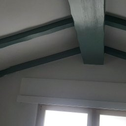 GONSKA DEVELOPMENT SPÓŁKA Z OGRANICZONĄ ODPOWIEDZIALNOŚCIĄ - Wnętrze poddasza w Gogolinie po odświeżeniu, z pomalowanymi na zielono drewnianymi belkami stropowymi i oknem w tle.