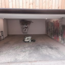 GONSKA DEVELOPMENT SPÓŁKA Z OGRANICZONĄ ODPOWIEDZIALNOŚCIĄ - Garaż po szpachlowaniu i malowaniu, zabezpieczone folią narty i kije narciarskie, odkurzacz przemysłowy na środku, surowe drewno na zewnątrz.