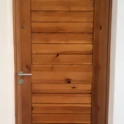 Drewniane drzwi wejściowe z poziomymi panelami, srebrną klamką i listwą progową, osadzone w białej framudze.