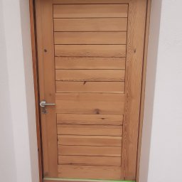 GONSKA DEVELOPMENT SPÓŁKA Z OGRANICZONĄ ODPOWIEDZIALNOŚCIĄ - Drewniane drzwi wewnętrzne z poziomymi panelami, zabezpieczone taśmą malarską i papierem przed malowaniem ścian. Widoczny pędzel i wiadro farby.