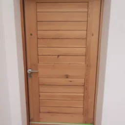 Drewniane drzwi wewnętrzne z poziomymi panelami, zabezpieczone taśmą malarską i papierem przed malowaniem ścian. Widoczny pędzel i wiadro farby.