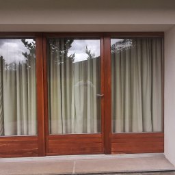 GONSKA DEVELOPMENT SPÓŁKA Z OGRANICZONĄ ODPOWIEDZIALNOŚCIĄ - Trzyczęściowe okno z drewnianą ramą i zasłonami w białym, otynkowanym budynku, oświetlone dwoma zewnętrznymi lampami.