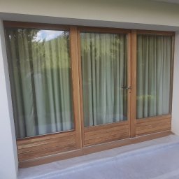 GONSKA DEVELOPMENT SPÓŁKA Z OGRANICZONĄ ODPOWIEDZIALNOŚCIĄ - Świeżo pomalowana, biała elewacja domu z drewnianymi drzwiami balkonowymi, zabezpieczona folią malarską, z widocznym wiadrem z farbą i drabiną.