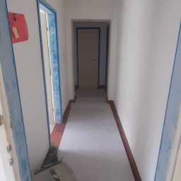 GONSKA DEVELOPMENT SPÓŁKA Z OGRANICZONĄ ODPOWIEDZIALNOŚCIĄ - Korytarz w trakcie zabezpieczania folią malarską, widoczny agregat szlifierski przy ścianie, przygotowanie do prac wykończeniowych.