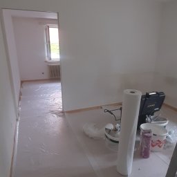 GONSKA DEVELOPMENT SPÓŁKA Z OGRANICZONĄ ODPOWIEDZIALNOŚCIĄ - Przygotowane do malowania pomieszczenie z zabezpieczoną folią podłogą, widoczne narzędzia malarskie, drabina i wiaderka z farbą.