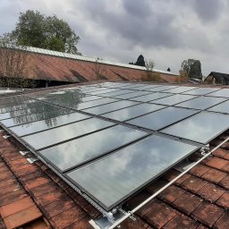 GONSKA DEVELOPMENT SPÓŁKA Z OGRANICZONĄ ODPOWIEDZIALNOŚCIĄ - Montaż paneli słonecznych na dachu z czerwonej dachówki, widoczne mocowania i odbicie chmur na panelach.