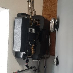 GONSKA DEVELOPMENT SPÓŁKA Z OGRANICZONĄ ODPOWIEDZIALNOŚCIĄ - Zamontowany, nowoczesny, czarny piec grzewczy z podłączonymi rurami miedzianymi, obok szary zbiornik i biały zbiornik wyrównawczy, widoczne także plastikowe pojemniki z płynem.