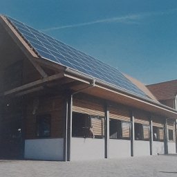 GONSKA DEVELOPMENT SPÓŁKA Z OGRANICZONĄ ODPOWIEDZIALNOŚCIĄ - Nowoczesna stajnia z panelami słonecznymi na dachu, drewniane ściany, białe fundamenty, błękitne niebo w tle.