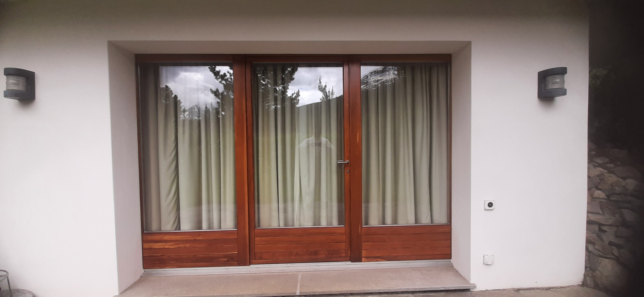 Trzyczęściowe okno z drewnianą ramą i zasłonami w białym, otynkowanym budynku, oświetlone dwoma zewnętrznymi lampami.