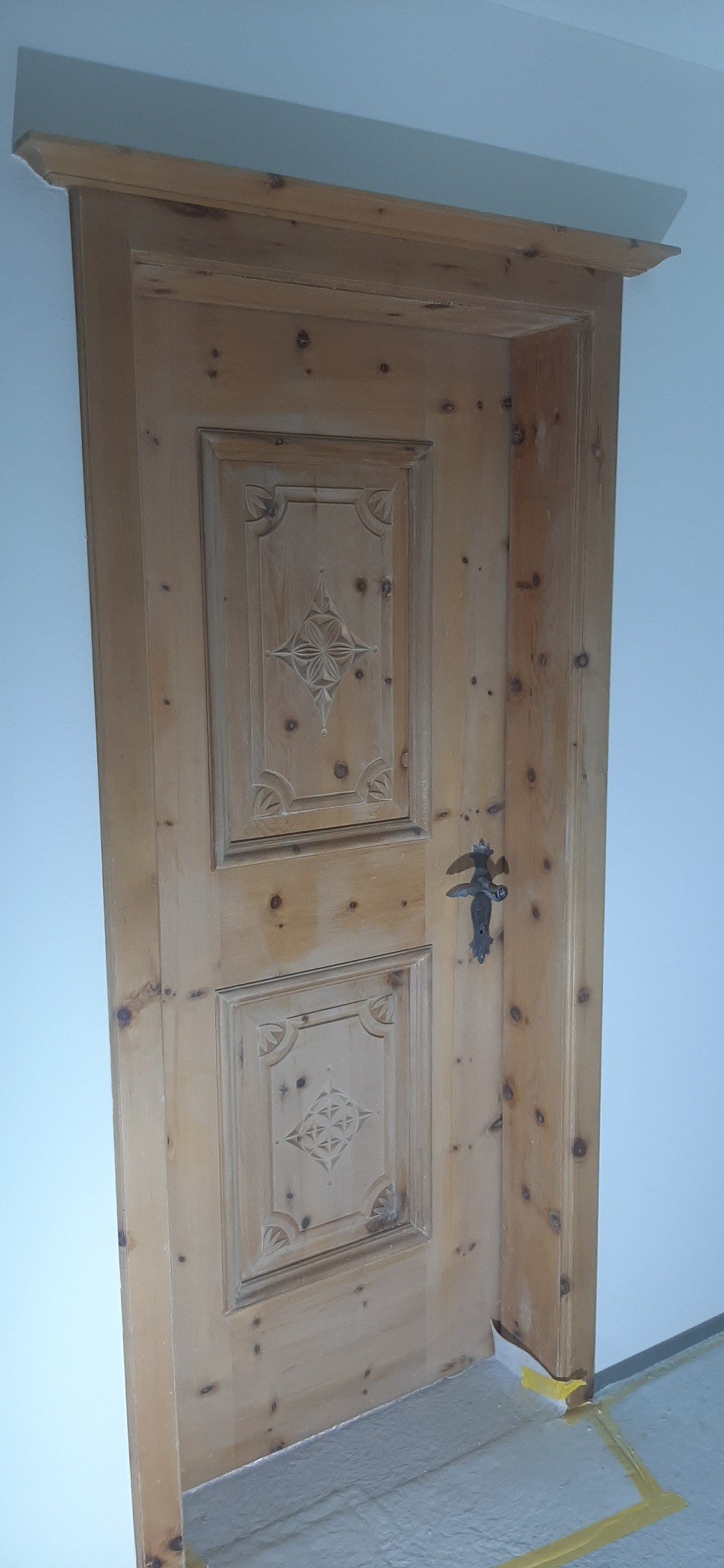 Odnowione drewniane drzwi wewnętrzne z ozdobnymi płaskorzeźbami i metalową klamką, zabezpieczone taśmą malarską przy podłodze.