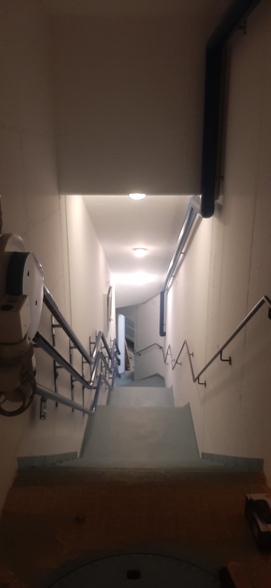 Klatka schodowa w piwnicy z metalowymi rurami i poręczami, widok z góry w dół, oświetlona lampami sufitowymi.