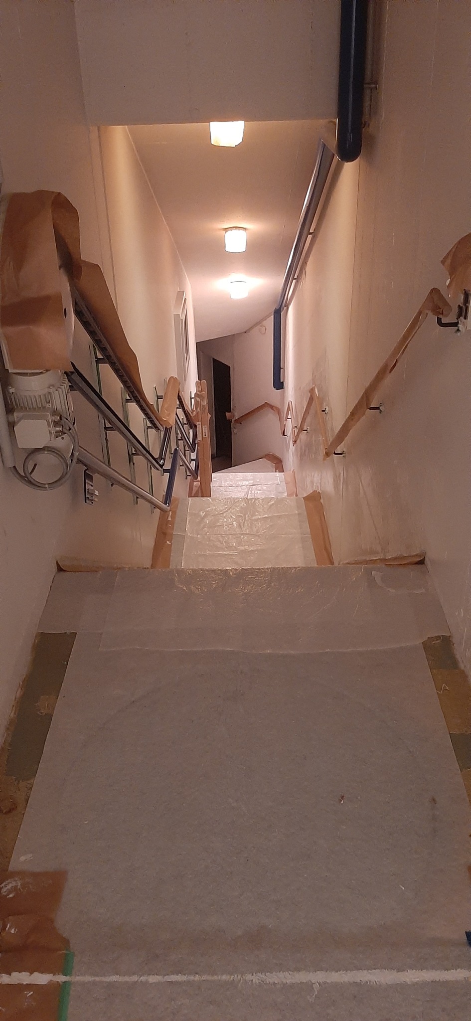 Klatka schodowa zabezpieczona folią i taśmą malarską przed pracami remontowymi, widok z góry na dół, z widocznymi elementami instalacji i schodami pokrytymi ochroną.