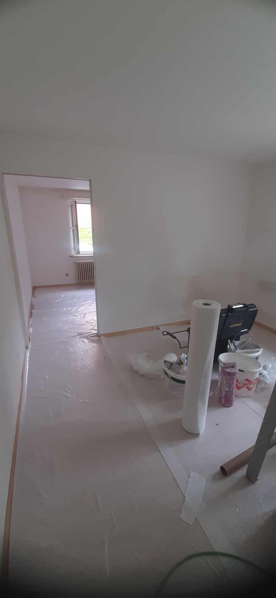 Przygotowane do malowania pomieszczenie z zabezpieczoną folią podłogą, widoczne narzędzia malarskie, drabina i wiaderka z farbą.