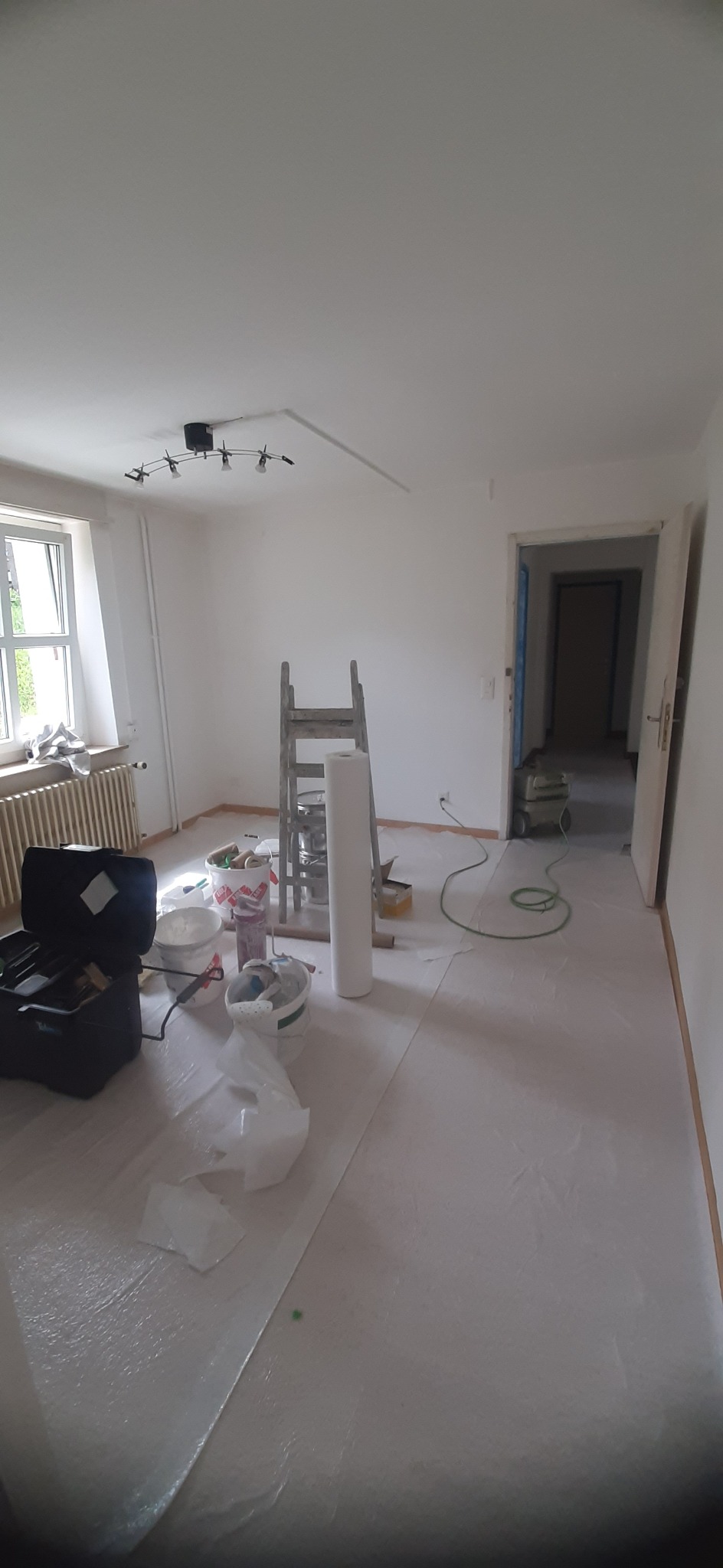 Pokój w trakcie przygotowań do odświeżenia: podłoga zabezpieczona folią ochronną, widoczne pędzle, wałki malarskie, drabina i wiaderka z farbą, ściany świeżo pomalowane na biało, w tle odkurzacz...