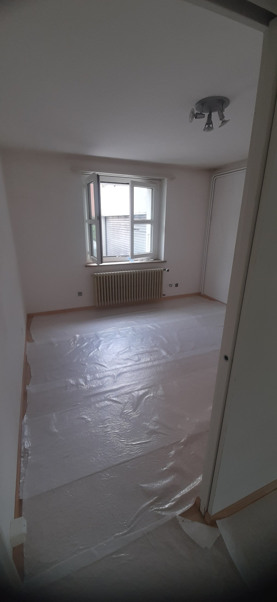 Pokój przygotowany do prac wykończeniowych, podłoga zabezpieczona folią malarską, ściany pomalowane na biało, okno otwarte, widoczny grzejnik i lampa sufitowa.