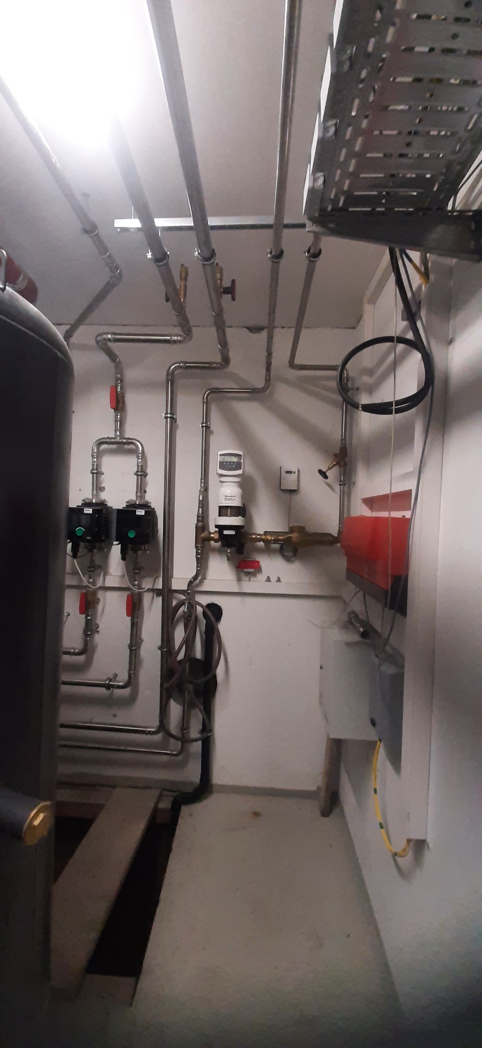 Pomieszczenie kotłowni z widocznymi elementami instalacji grzewczej: rury, zawory, pompy obiegowe, zbiornik akumulacyjny i skrzynka elektryczna.