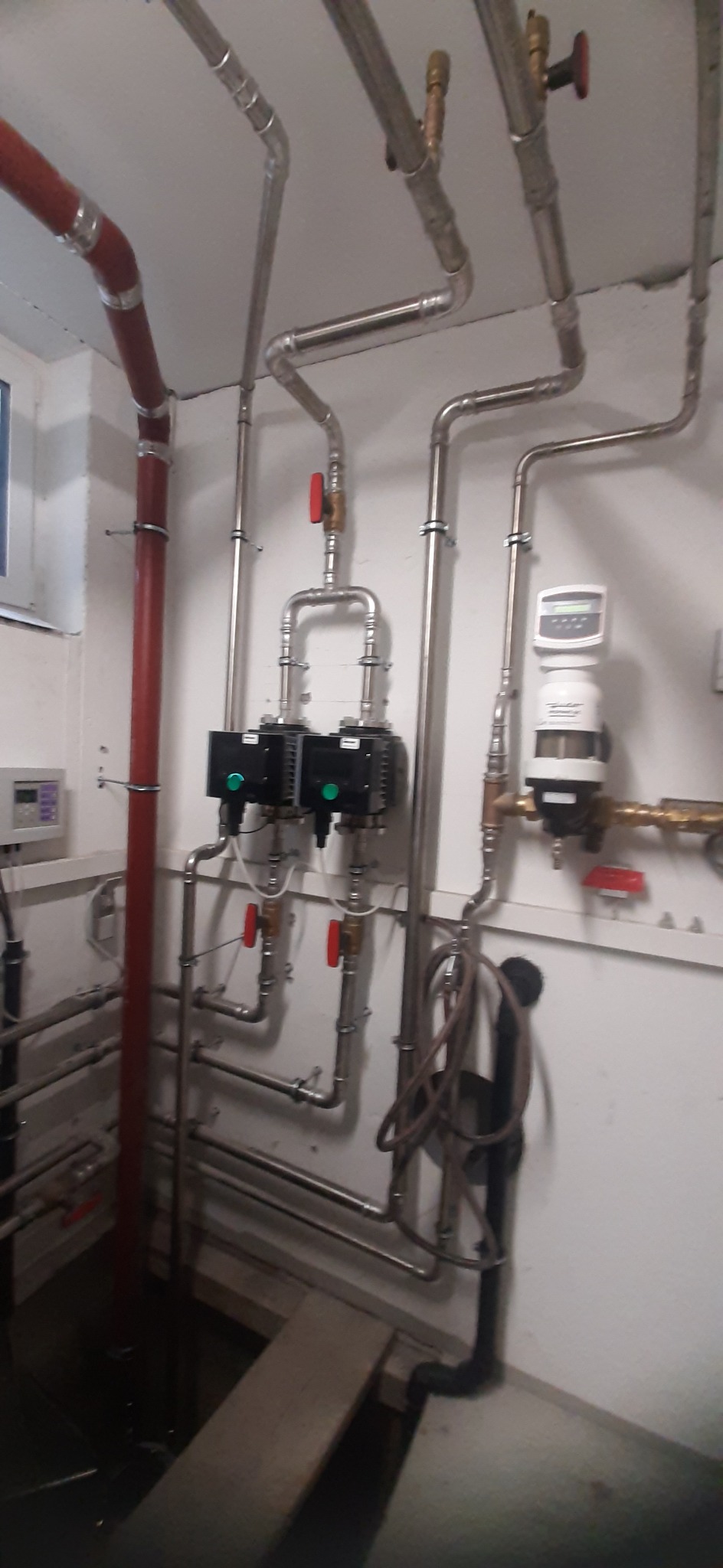 Węzeł cieplny w piwnicy z rurami miedzianymi i stalowymi, pompami obiegowymi i licznikiem ciepła na białej ścianie.