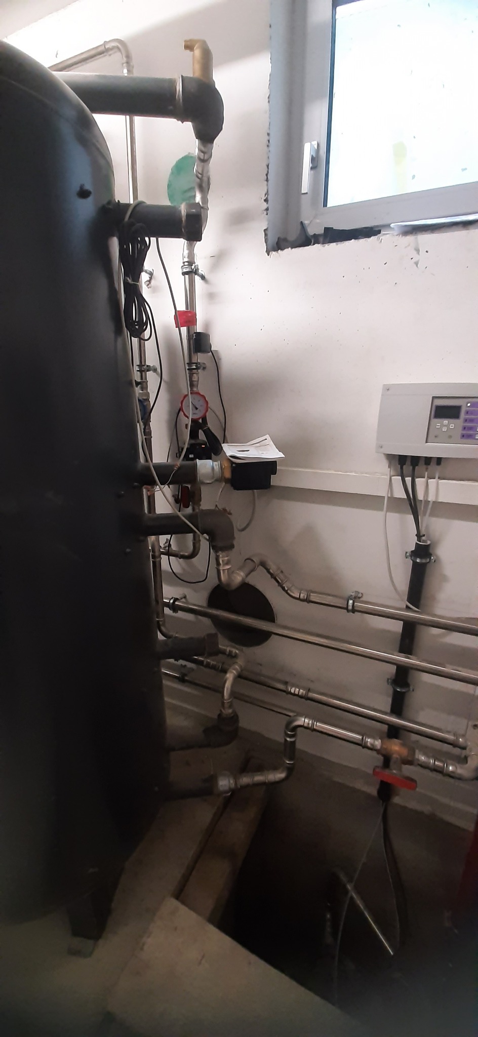 Czarny zbiornik buforowy z podłączonymi rurami i manometrem w kotłowni, widoczne okno i sterownik na ścianie.