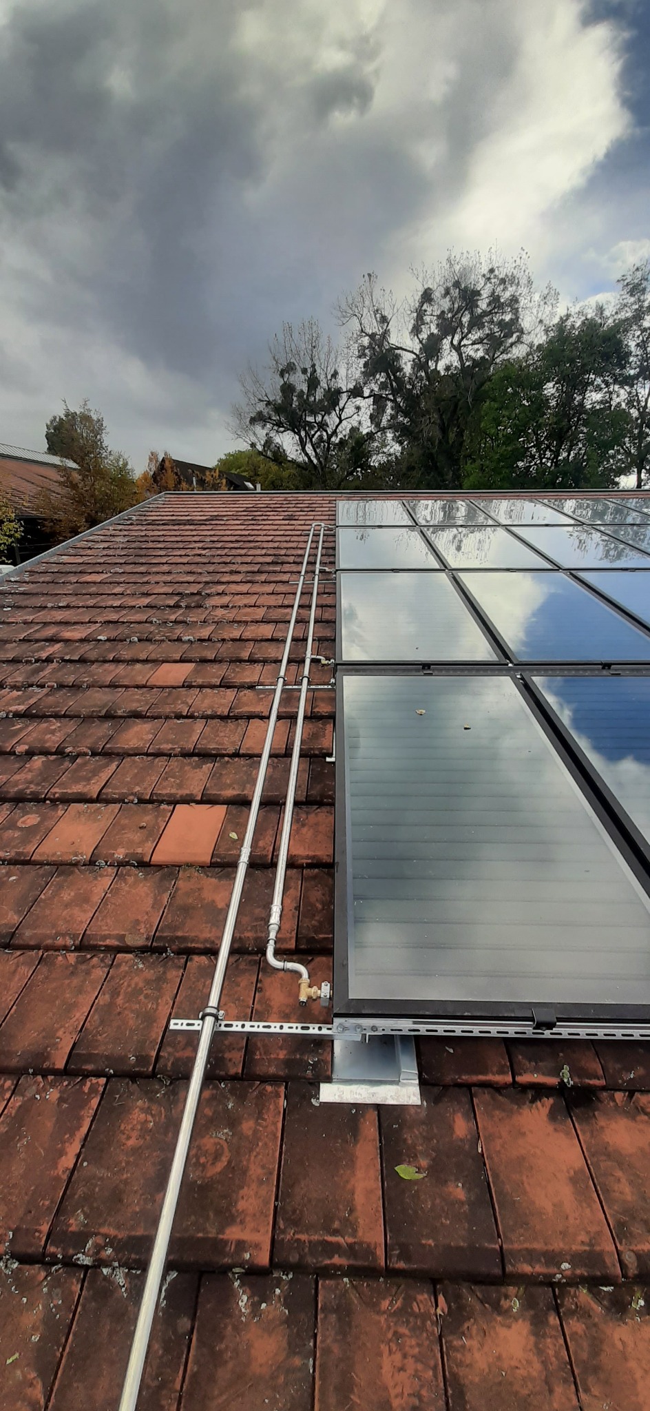 Panele solarne zamontowane na pomarańczowej dachówce pod pochmurnym niebem, widoczne rury instalacyjne i elementy mocujące.