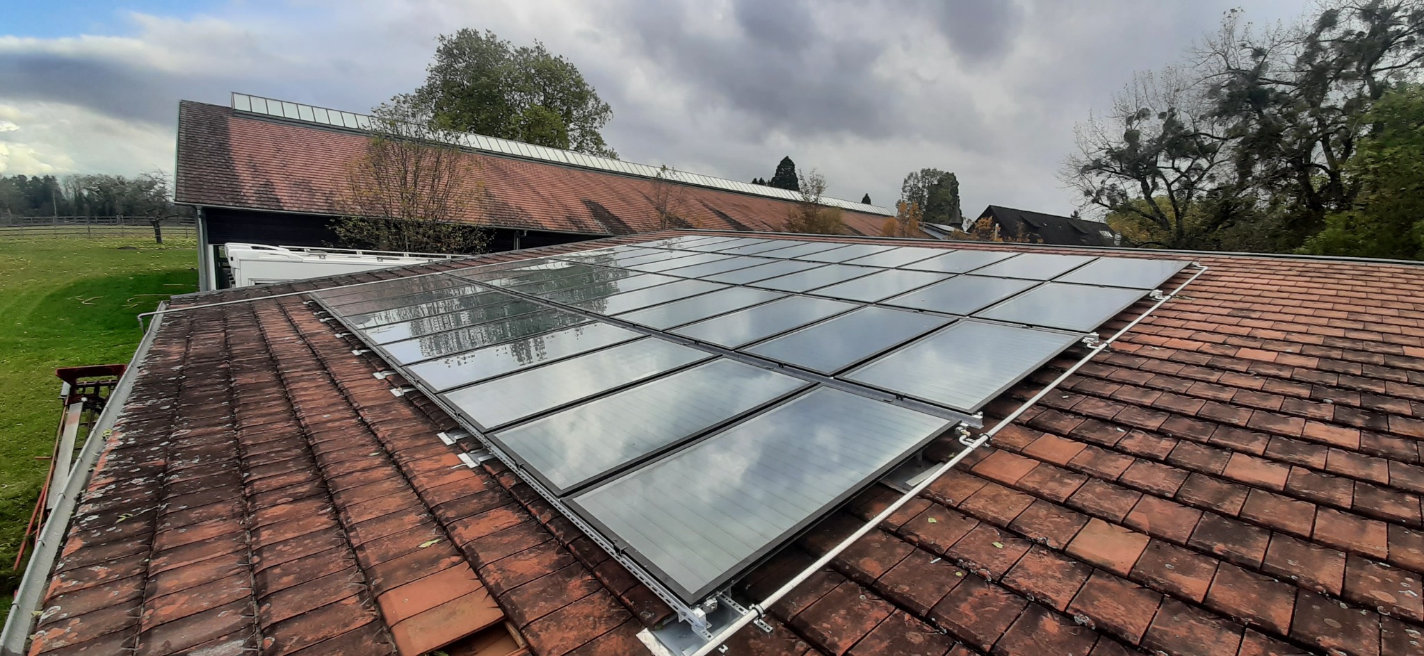 Montaż paneli słonecznych na dachu z czerwonej dachówki, widoczne mocowania i odbicie chmur na panelach.