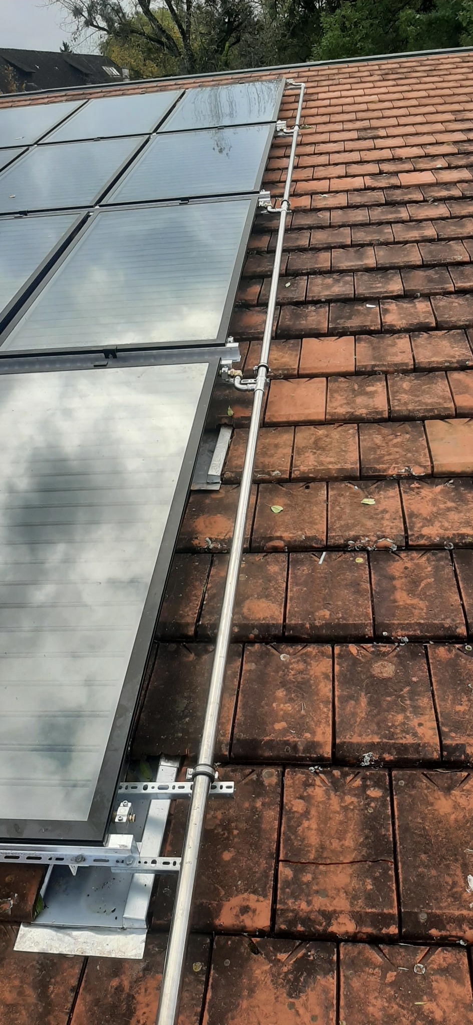 Montaż paneli słonecznych na dachu z czerwonej dachówki, widoczne metalowe wsporniki i rury instalacyjne.