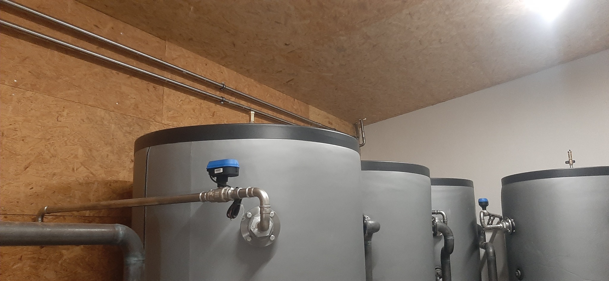 Rząd szarych, cylindrycznych zbiorników z rurami i czujnikami w pomieszczeniu z boazerią i białymi ścianami.