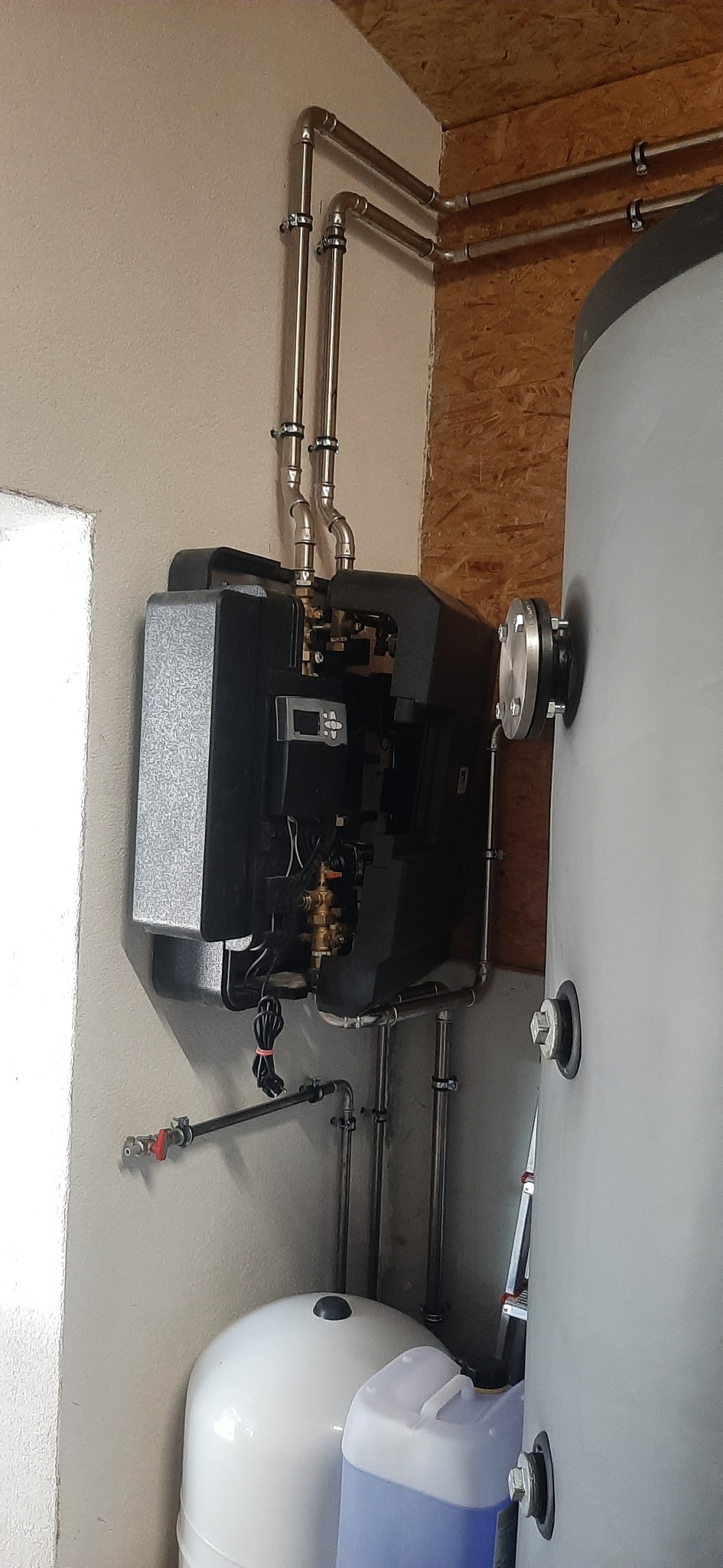 Zamontowany, nowoczesny, czarny piec grzewczy z podłączonymi rurami miedzianymi, obok szary zbiornik i biały zbiornik wyrównawczy, widoczne także plastikowe pojemniki z płynem.