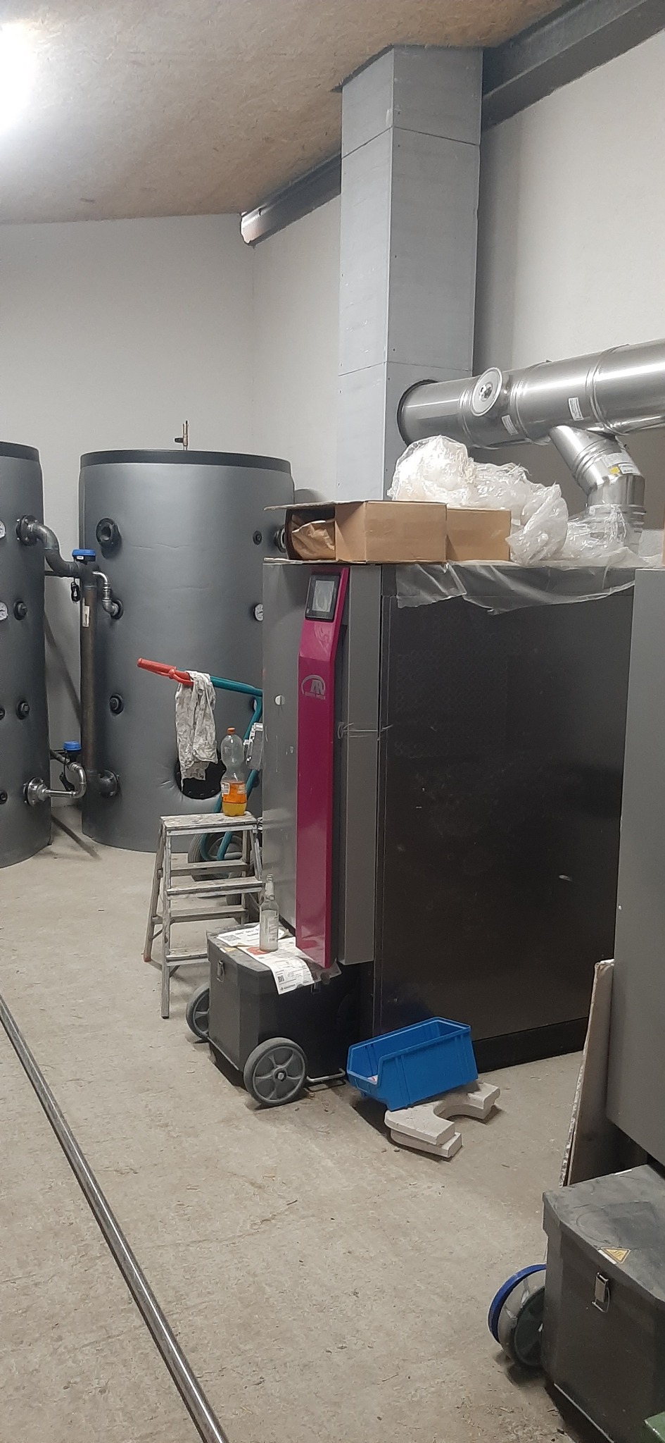Wnętrze kotłowni z dwoma szarymi zbiornikami buforowymi, kotłem na paliwo stałe z bordowym panelem i metalową rurą wentylacyjną pod sufitem. Na kotle leżą kartony i folie, a obok stoją drabina...