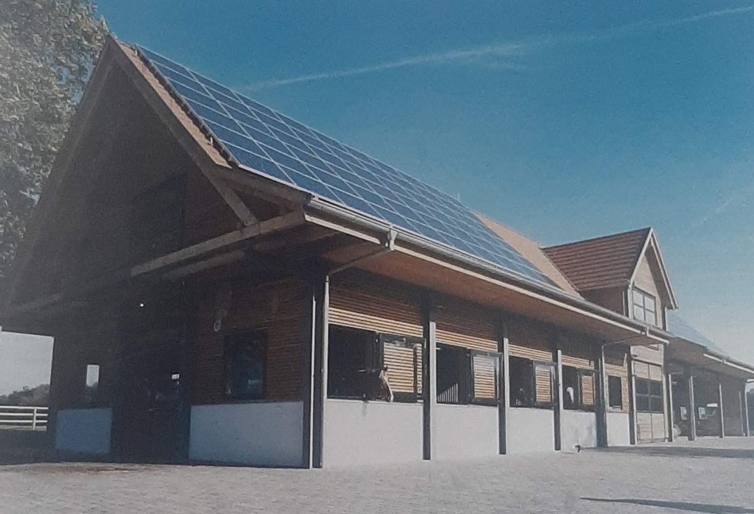 Nowoczesna stajnia z panelami słonecznymi na dachu, drewniane ściany, białe fundamenty, błękitne niebo w tle.