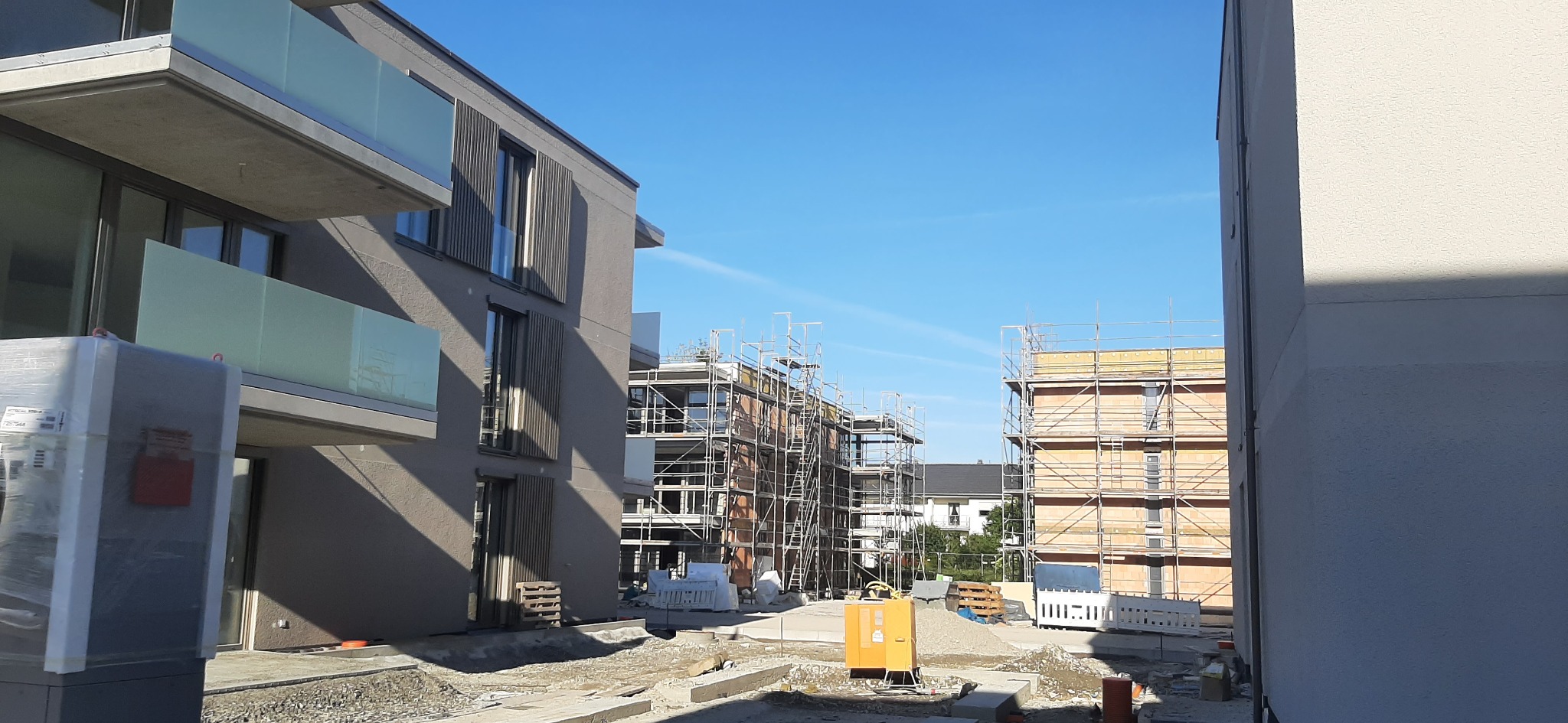 Nowe budynki w trakcie wykańczania elewacji, widoczne rusztowania, przygotowane materiały budowlane i żółty agregat prądotwórczy.