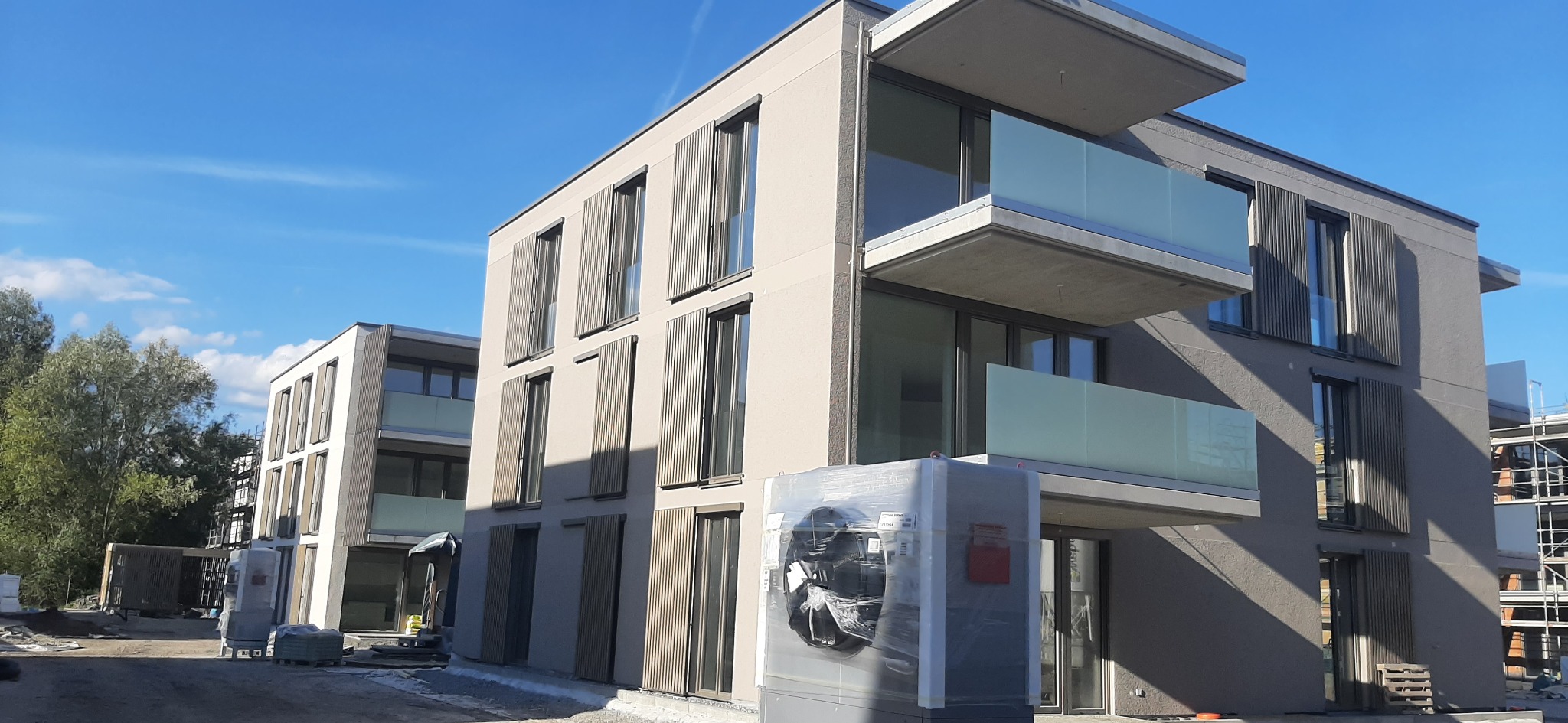 Dwa nowoczesne budynki wielorodzinne z szarymi elewacjami i drewnianymi żaluzjami okiennymi, w tle błękitne niebo, a na pierwszym planie zapakowana jednostka klimatyzacyjna.