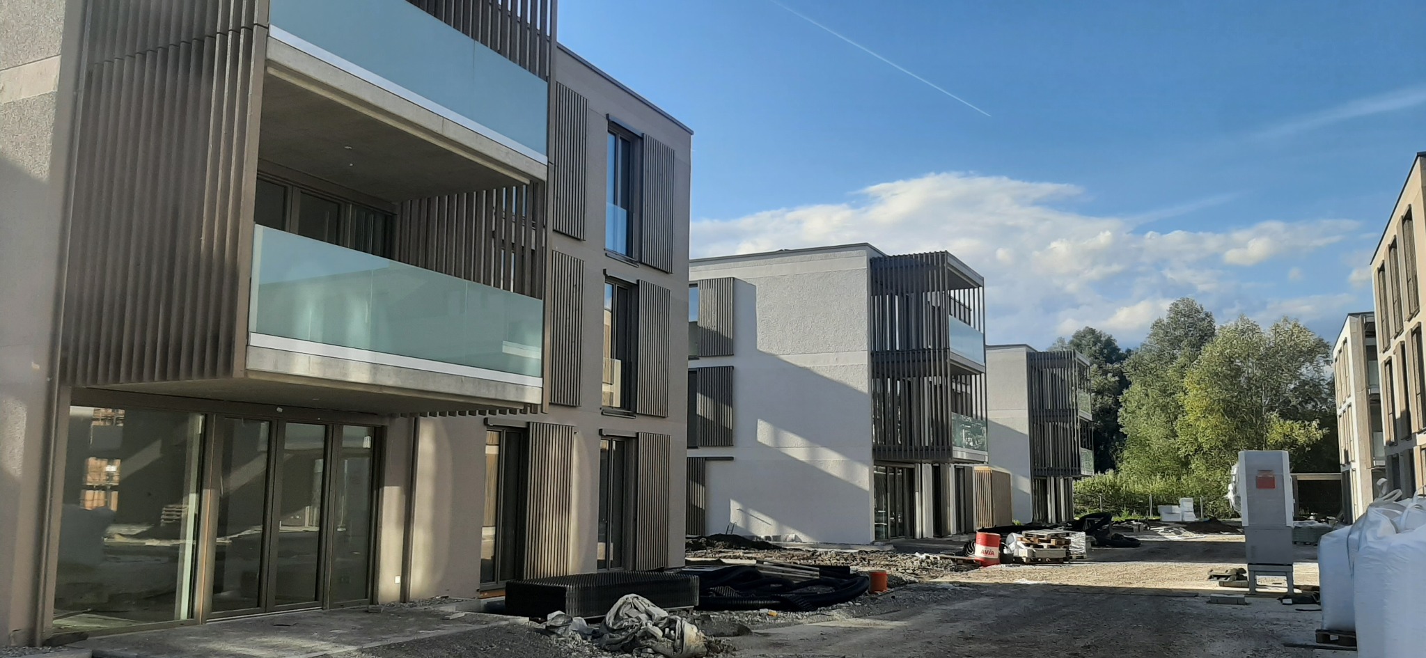 Nowoczesne, wielorodzinne budynki z balkonami o szklanych balustradach i drewnianymi żaluzjami, w trakcie prac wykończeniowych na zewnątrz, z widocznymi materiałami budowlanymi i lekkim...