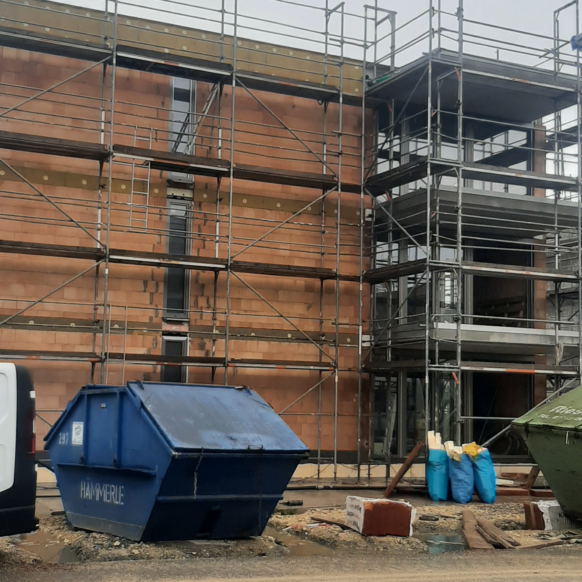 Budynek w trakcie ocieplania elewacji, widoczne rusztowania, warstwa izolacyjna na ścianie, niebieski kontener na odpady budowlane z napisem HAMMERLE, obok zaparkowany biały bus.