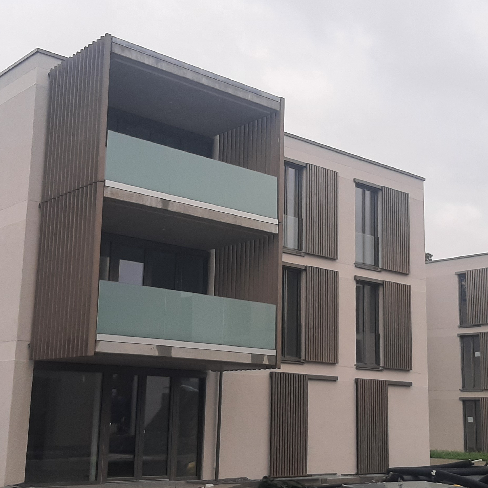 Nowoczesny budynek wielorodzinny z balkonami wykończonymi szkłem i drewnianymi lamelami, widoczne rury na dole elewacji.