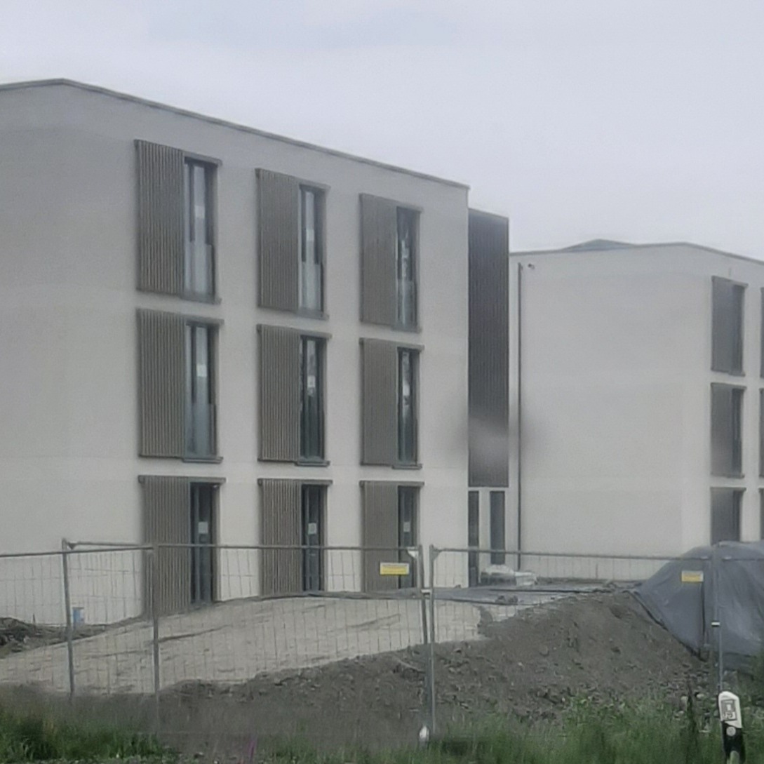 Nowoczesny budynek wielorodzinny z jasną elewacją i drewnianymi żaluzjami okiennymi w trakcie budowy, ogrodzony metalowym płotem.