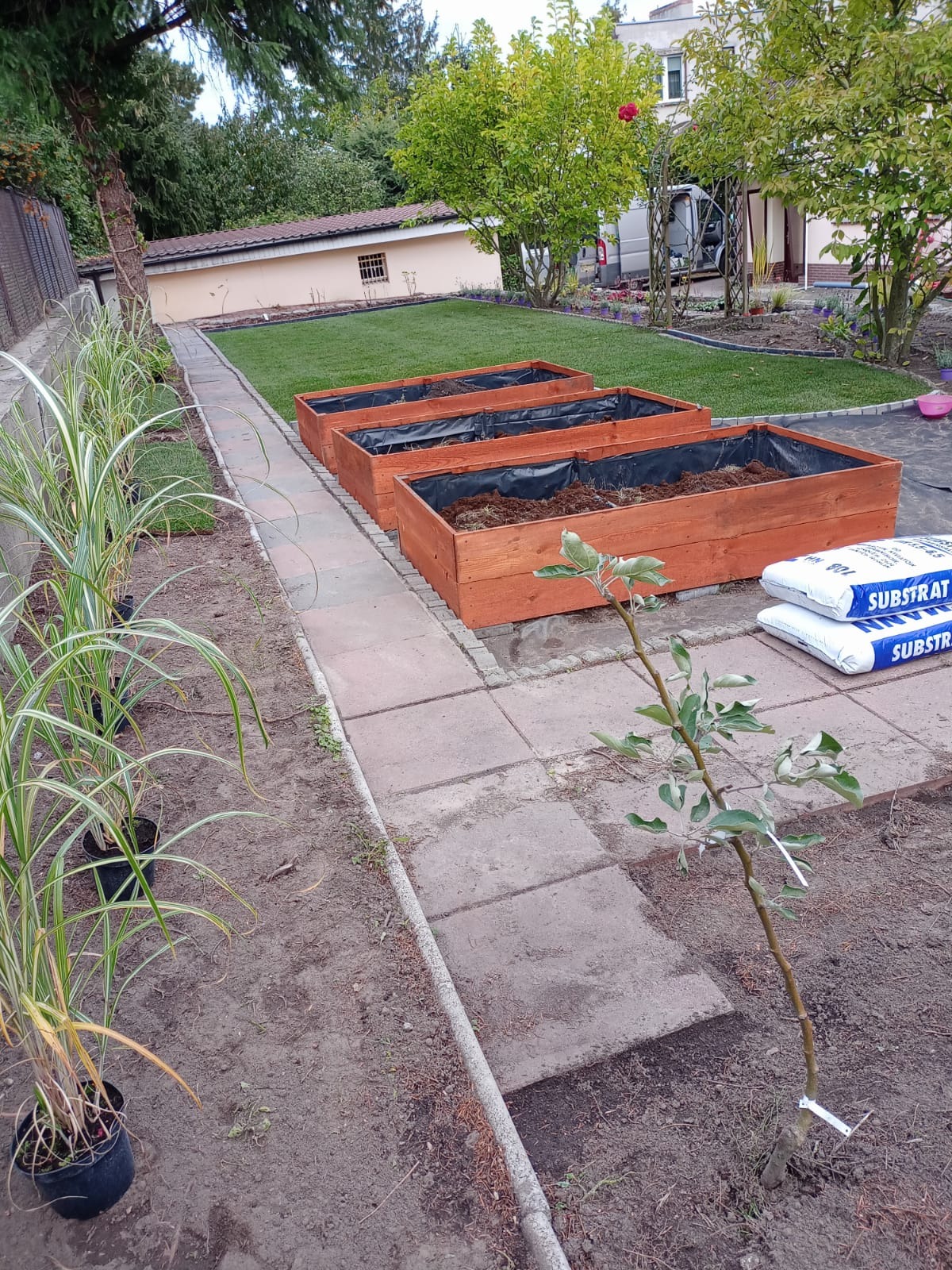 Przygotowanie do założenia ogrodu: trzy drewniane skrzynie wyłożone folią i wypełnione ziemią, worki z substratem obok, sadzonki w doniczkach i świeżo posadzone drzewko owocowe na tle trawnika...