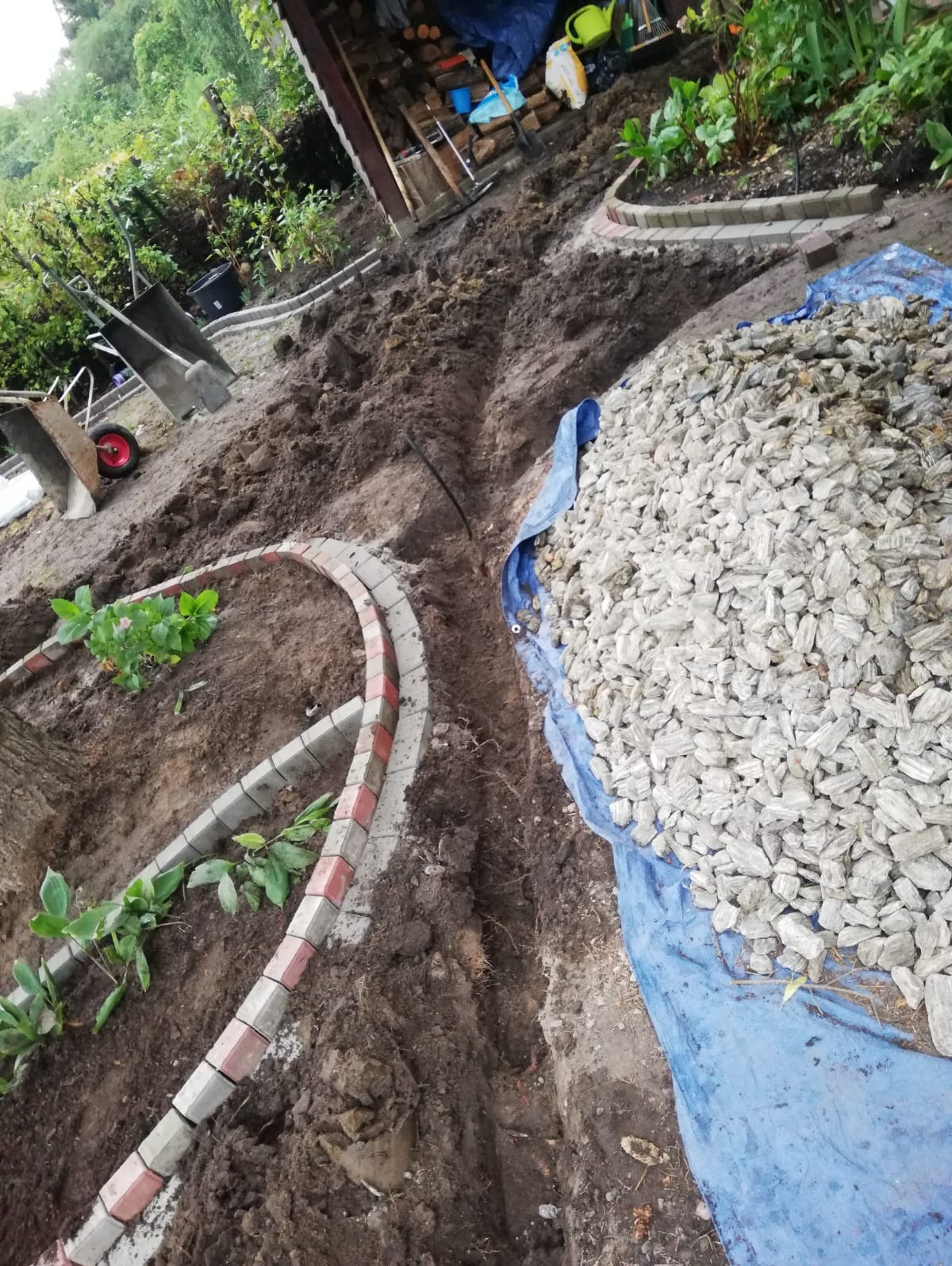 Przygotowanie terenu pod ścieżkę w ogrodzie: wykopany rów, ułożone obrzeża z kostki i pryzma kamieni na niebieskiej plandece.