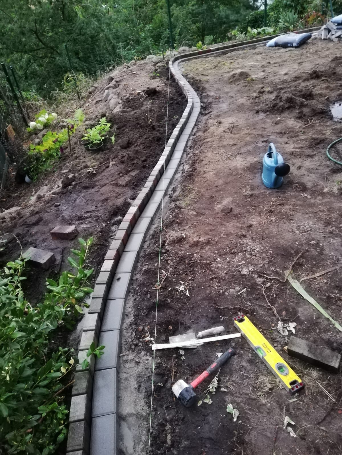 Wykonywanie obrzeża z kostki brukowej w ogrodzie na nierównym terenie, widoczne narzędzia: poziomica, gumowy młotek, sznurek wyznaczający linię, niebieska konewka w tle.
