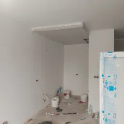 Wnętrze w trakcie remontu: ściany po gładziowaniu, przygotowane do malowania, widoczne puszki instalacyjne, sufit podwieszany w trakcie montażu, narzędzia i materiały budowlane porozrzucane...