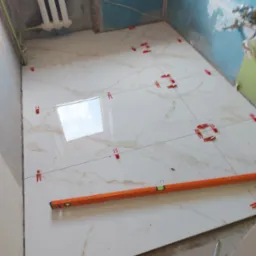 Podłoga w trakcie układania płytek ceramicznych imitujących marmur, z użyciem poziomicy i klipsów dystansowych, widoczne narzędzia glazurnicze i niedokończone ściany.