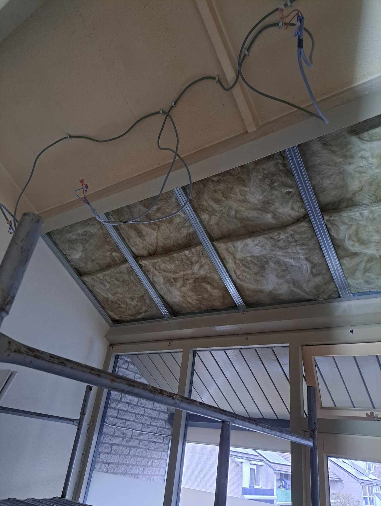 Izolacja termiczna poddasza z widoczną wełną mineralną między metalowymi profilami, kable elektryczne przymocowane do sufitu, widok z rusztowania na okno dachowe i fragment budynku na zewnątrz.